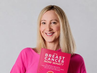 Christina Wilhelm is wearing a pink blouse and holding her book called 'It's Breast Cancer. Now What?' with the subtitle 'The missing manual for informed decisions, return to work and long-term recovery'