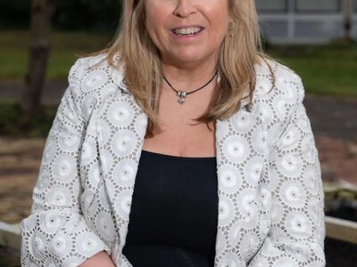 A headshot of Jill. She has blonde hair and is wearing black trousers with a black top and a white patterned blazer. She's sitting outside and smiling at the camera.