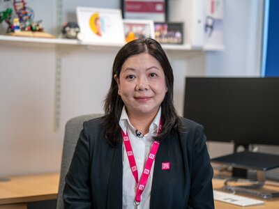 Dr Maggie Cheang, with dark hair, a blazer and pink BCN lanyard, posing for portrait photos in her office.