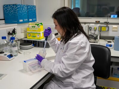Dr Maggie Cheang, with dark hair, in a white lab coat posing for portrait photos in a laboratory.