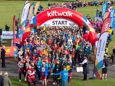 A crowd of participants passing under the inflatable arch at the start of Kiltwalk Edinburgh. They are wearing kilts and a man is playing the bagpipes.
