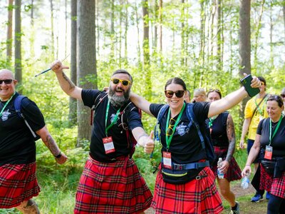 A group of smiling people walking through a wood as they take part in Kiltwalk Dundee. They are wearing kilts and black t-shirts.