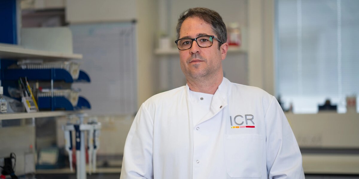 Issac, a man with short grey hair, black square frame glasses, in a white BCN lab coat, posing for portraits in a white lab coat with the lab in the background.