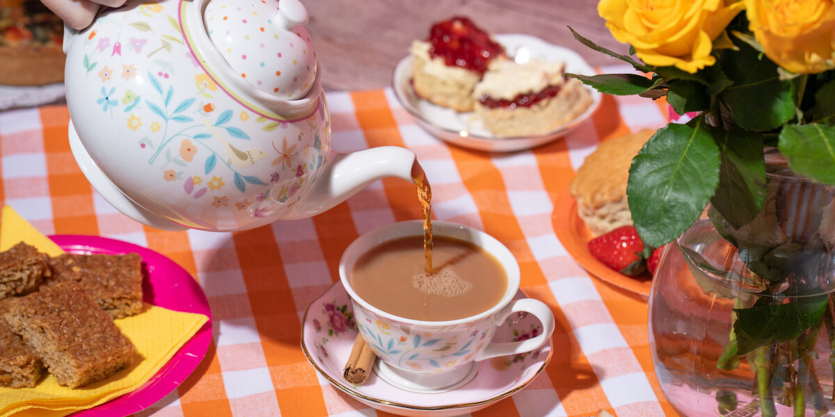 Photo of pouring tea from a teapot into a cup with a saucer at an Afternoon Tea event. Around the cup are plates of flapjack, victoria sponge and scones, as well as a vase of yellow flowers. They are all on a orange and white checked tablecloth.