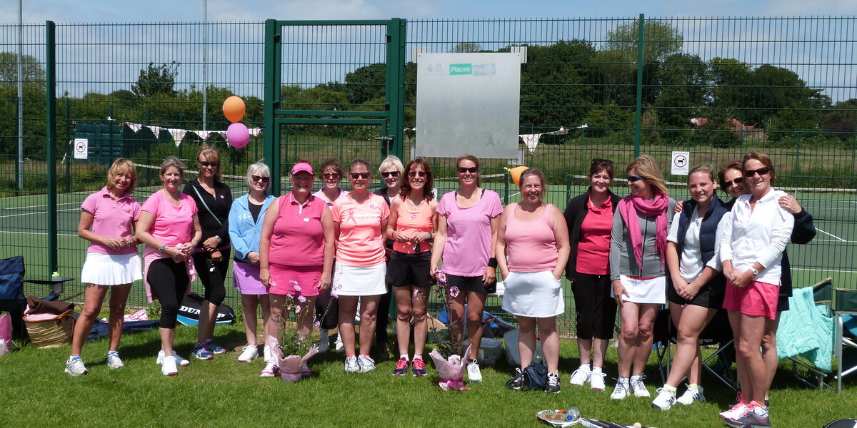 Group photo of 16 people standing smiling in front of tennis courts. Most are wearing sporty clothes in shades of pink. Tennis rackets are on the grass in the foreground.