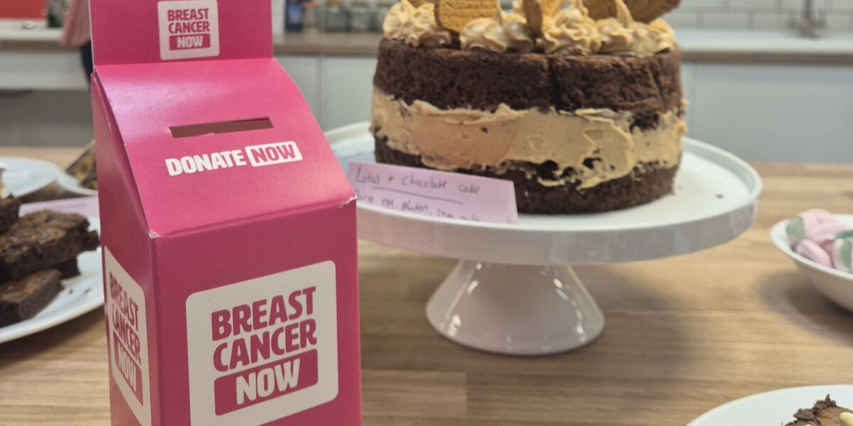 A photo of an Afternoon Tea donation box next to a cake on a cakestand. In the background is pink and white Breast Cancer Now bunting and someone wearing pink.