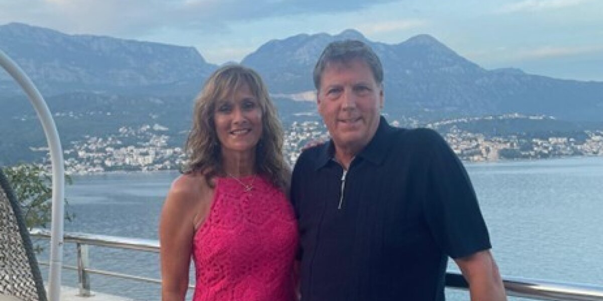 A photo of a couple standing on a balcony on holiday with a view of the ocean and mountains behind them. The woman is wearing a pink dress and the man is wearing pale blue shorts with a navy blue shirt.