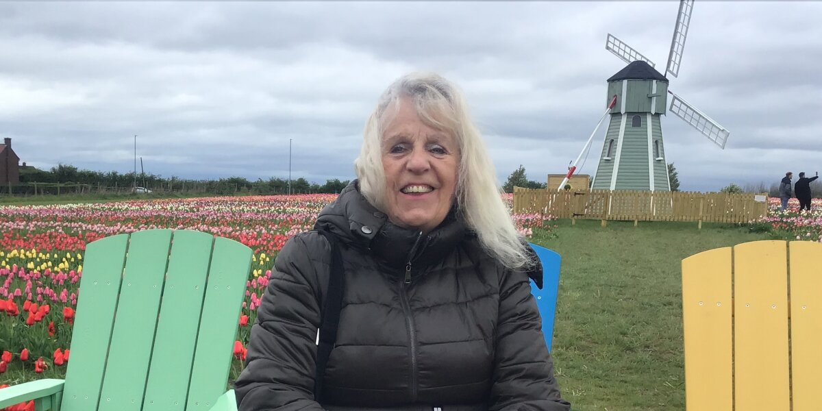 A women sitting on a wooden chair in front of colourful tulips in the Netherlands with a typical Dutch windmill in the background.