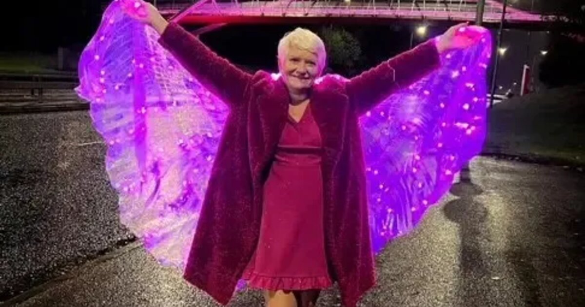 A photo of Julie wearing a pink dress, coat and shoes. She is smiling and has her arms outstretched holding pink light-up fairy wings. In the background a bridge is lit up pink.