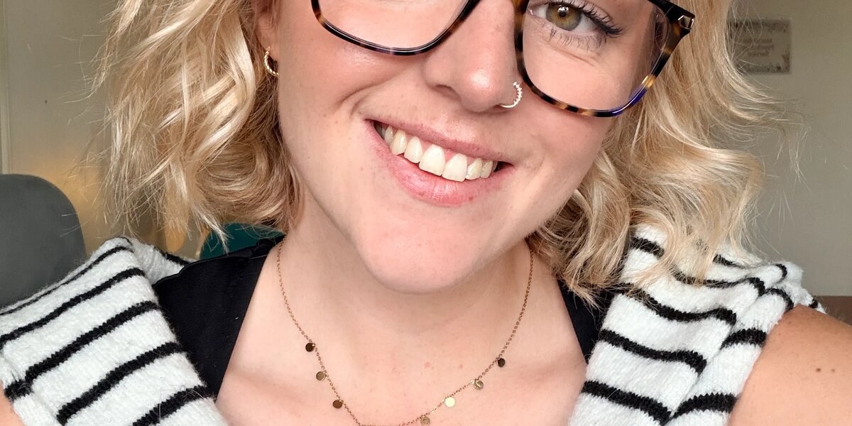 Selfie of a woman with blonde short hair and glasses. She is smiling at the camera wearing a black top with a black and white striped jumper tied around her shoulders.