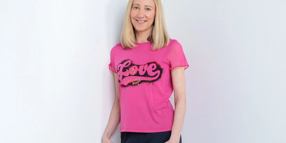 Christina has blonde hair. She's wearing a pink T-shirt with the word 'Love' on it with black leggings and pink high heels.