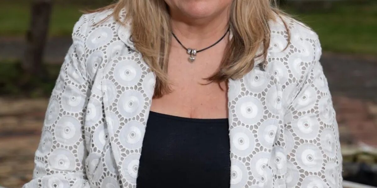 A headshot of Jill. She has blonde hair and is wearing black trousers with a black top and a white patterned blazer. She's sitting outside and smiling at the camera.