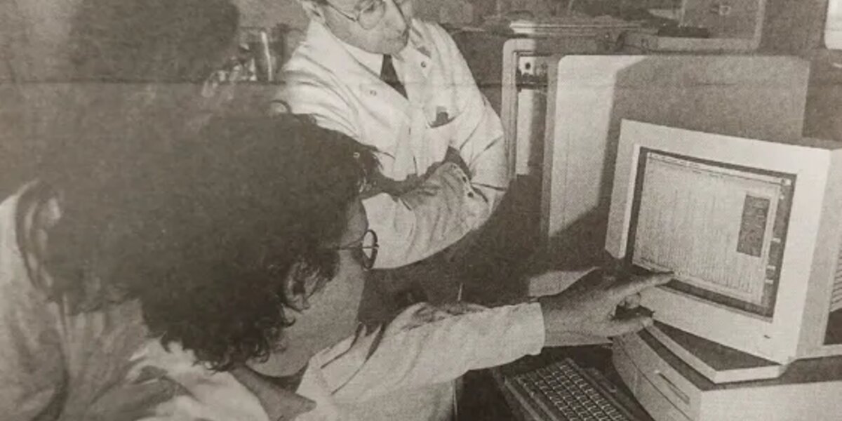 A black and white newspaper photo of Professor Mike Stratton and 2 other research colleagues gathered around a computer. They are all wearing lab coats and one is pointing to a graph on the computer monitor.