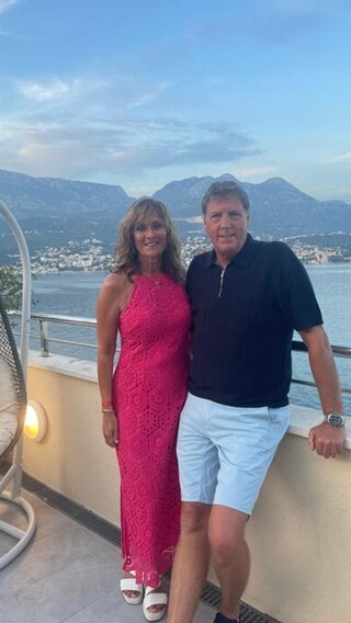 A photo of a couple standing on a balcony on holiday with a view of the ocean and mountains behind them. The woman is wearing a pink dress and the man is wearing pale blue shorts with a navy blue shirt.