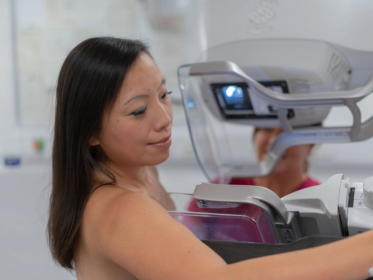 A mammographer in maroon scrubs, and a woman with dark hair demonstrating a mammogram screening.