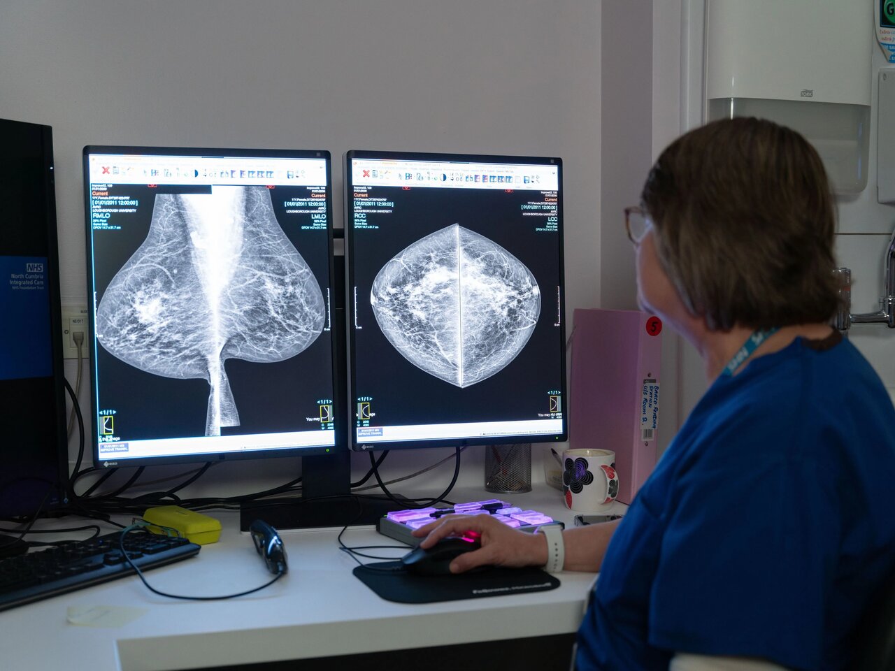 Karen, a consultant radiographer, with short dark hair and glasses, next to a computer screen in a dark room, looking at x-rays of breasts taken in the mammography suite.