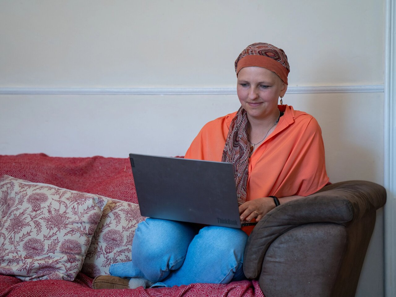 Claire, a woman in a bright orange top, with a red/brown leaf patterned headscarf and blue eyes, sat on her sofa, on her laptop.