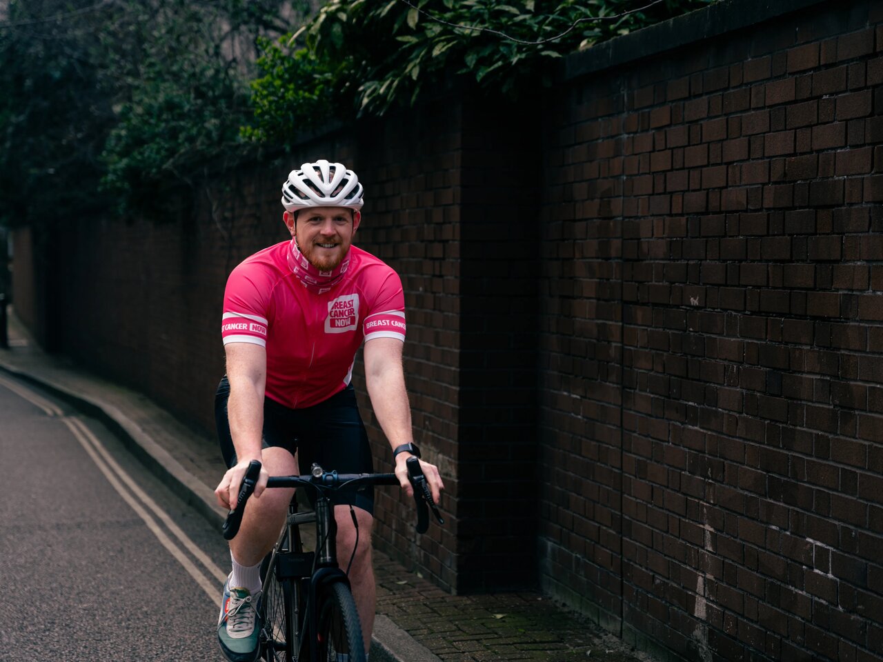 Breast Cancer Now staff wearing merchandise. Staff member Simon, wearing Breast Cancer Now cycling jersey, whilst outside on his bike.