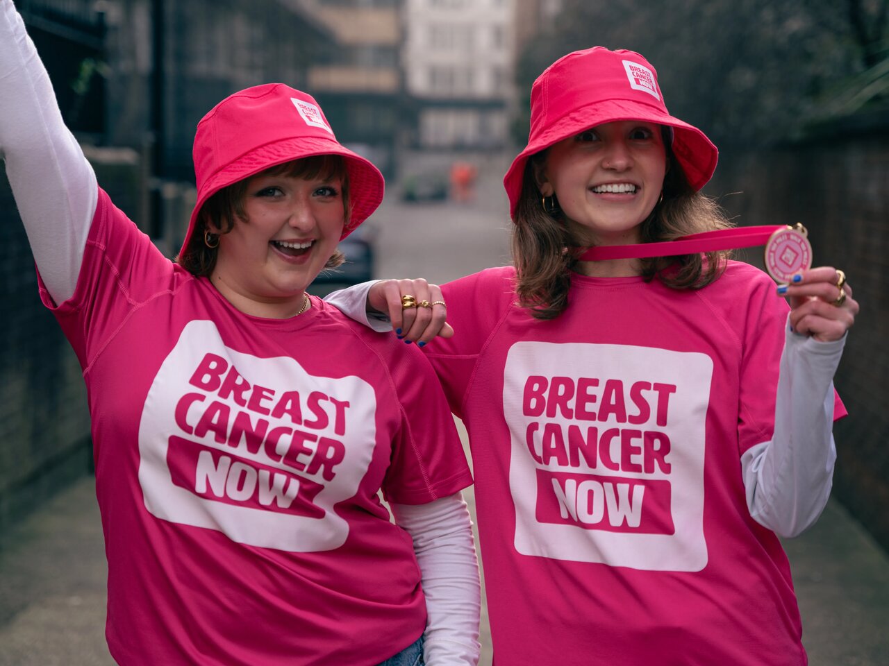 Two young women wearing pink bucket hats and cotton r-shirts with a white Breast Cancer Now logo, and a woodern medal on a ribbon, smiling. 