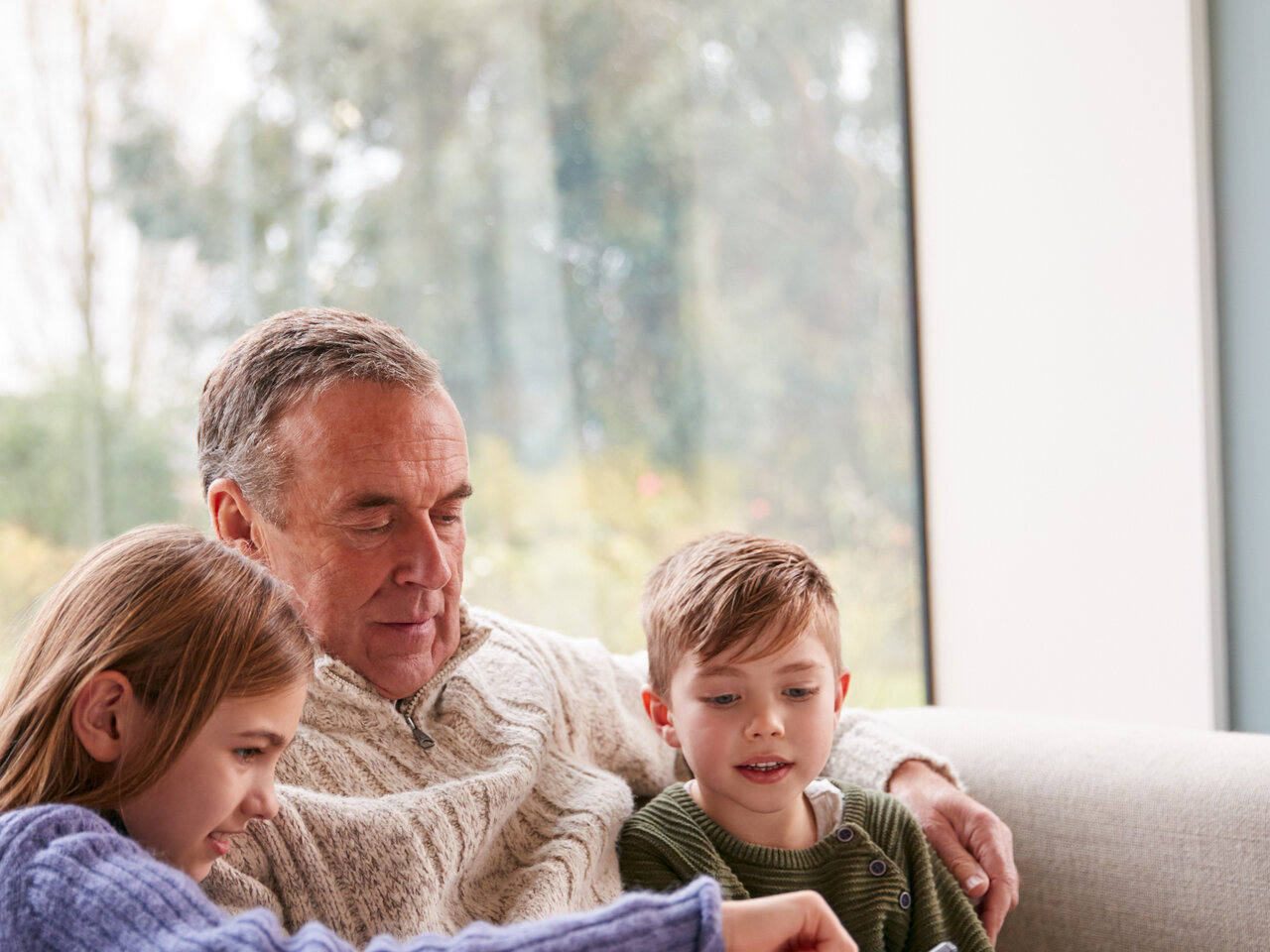 An older man sitting with two young children on a sofa. They're all looking at something on a mobile phone.