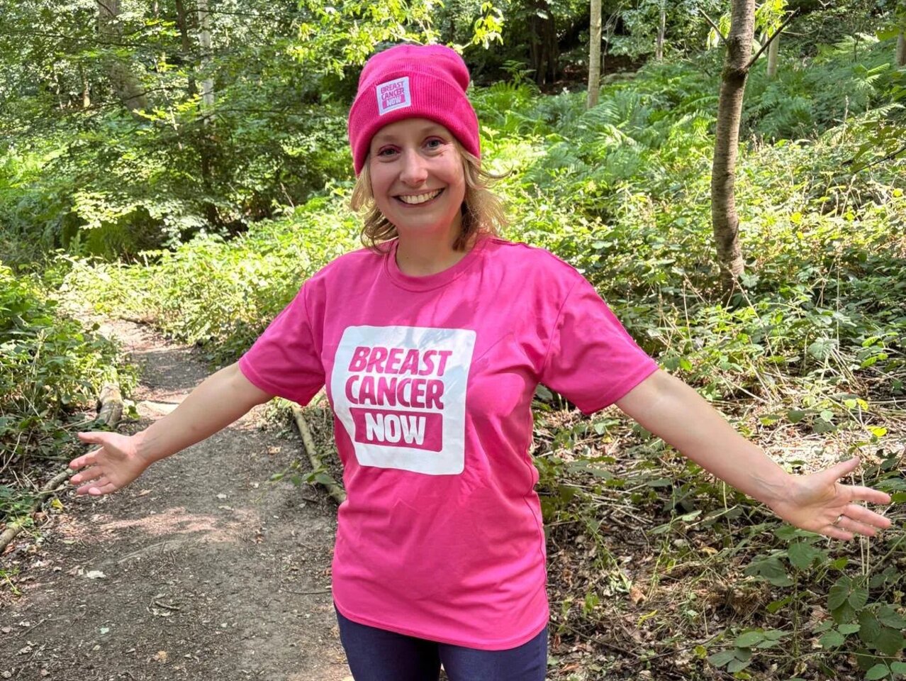 Georgie is standing on a path surrounded by woodland on a sunny day. She is wearing jeans and her pink Breast Cancer Now bucket hat and T-shirt. She is smiling at the camera.