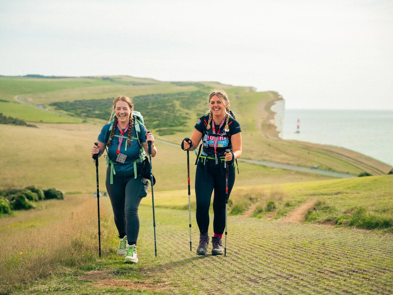 Photo of 2 women with outdoor gear and walking poles taking part in the South Coast Ultra Challenge. In the background is countryside and chalk cliffs by the sea.
