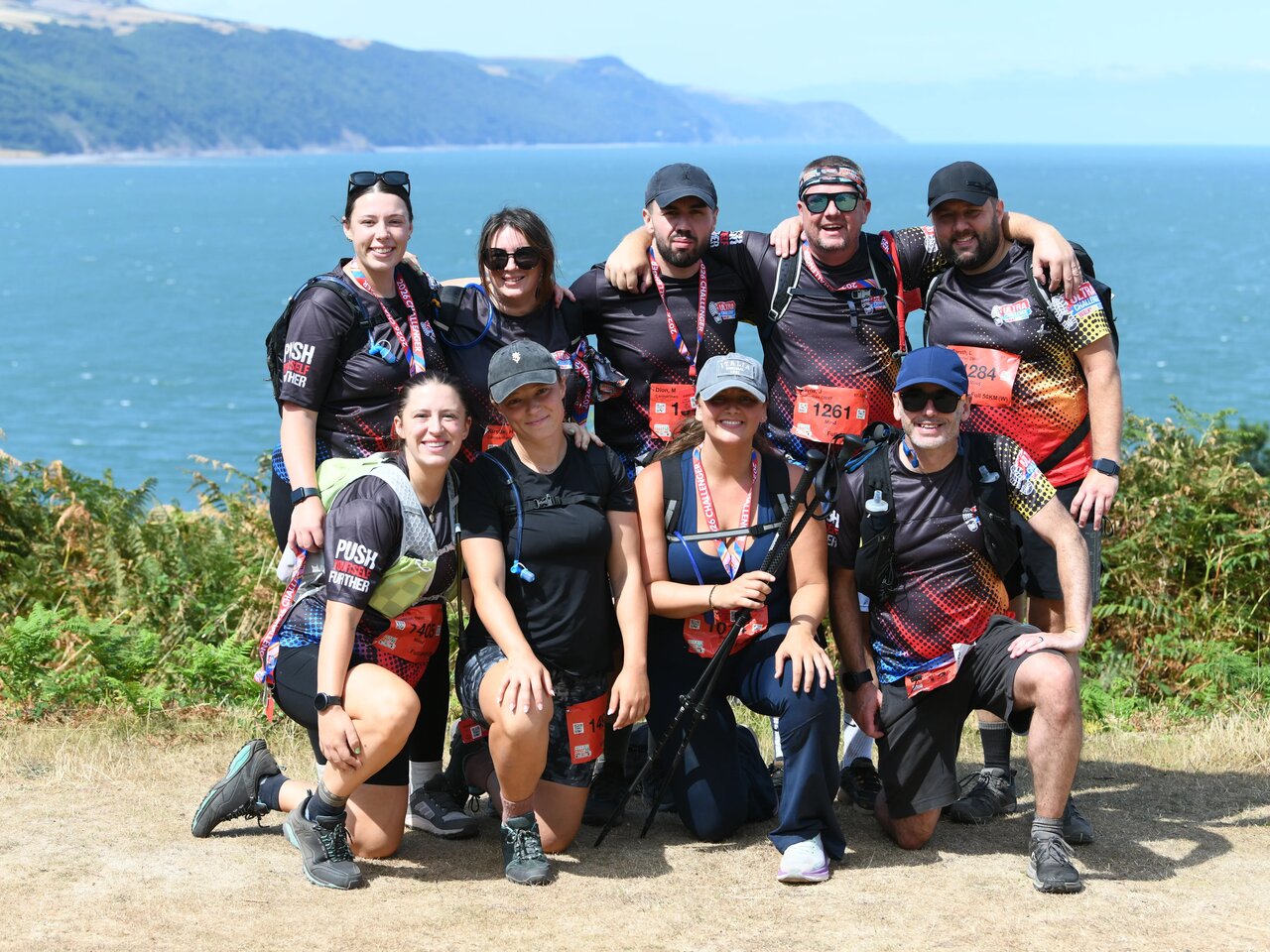 Group photo of 9 people taking part in the South West Coast 50 Ultra Challenge on a sunny day. They are wearing walking gear and smiling. In the background the sea and coast are visible.