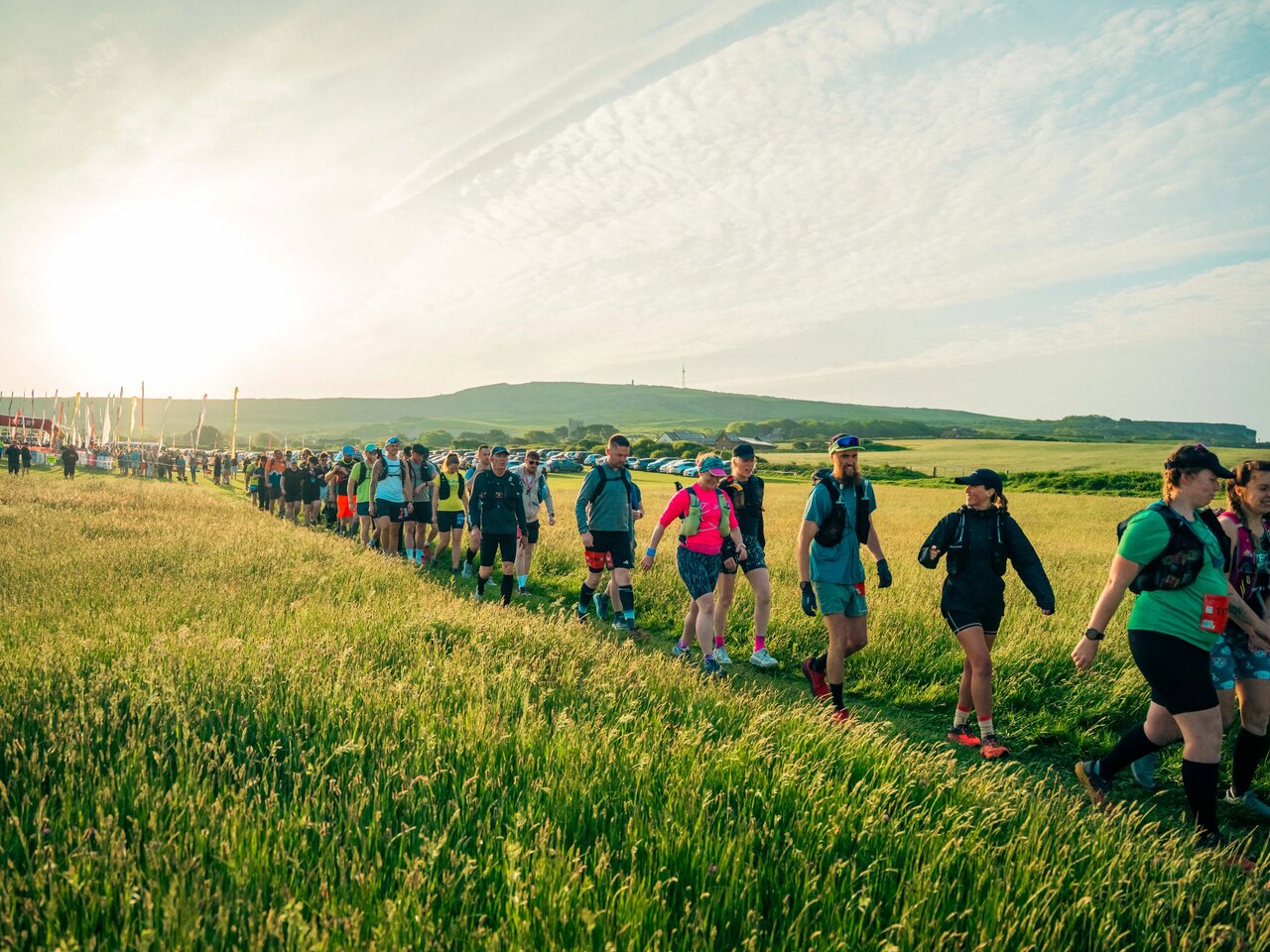 Photo of a long line of people walking through a sunny field as they begin the Isle of Wight Ultra Challenge. The start line and flags can be seen in the background.