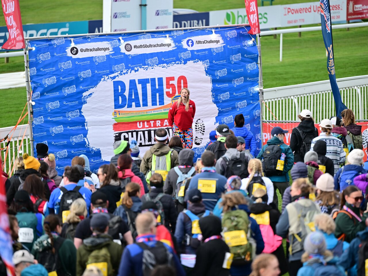 Photo of a woman on an outdoor stage at a racecourse standing in front of a large group of participants at the Bath 50 Ultra Challenge.