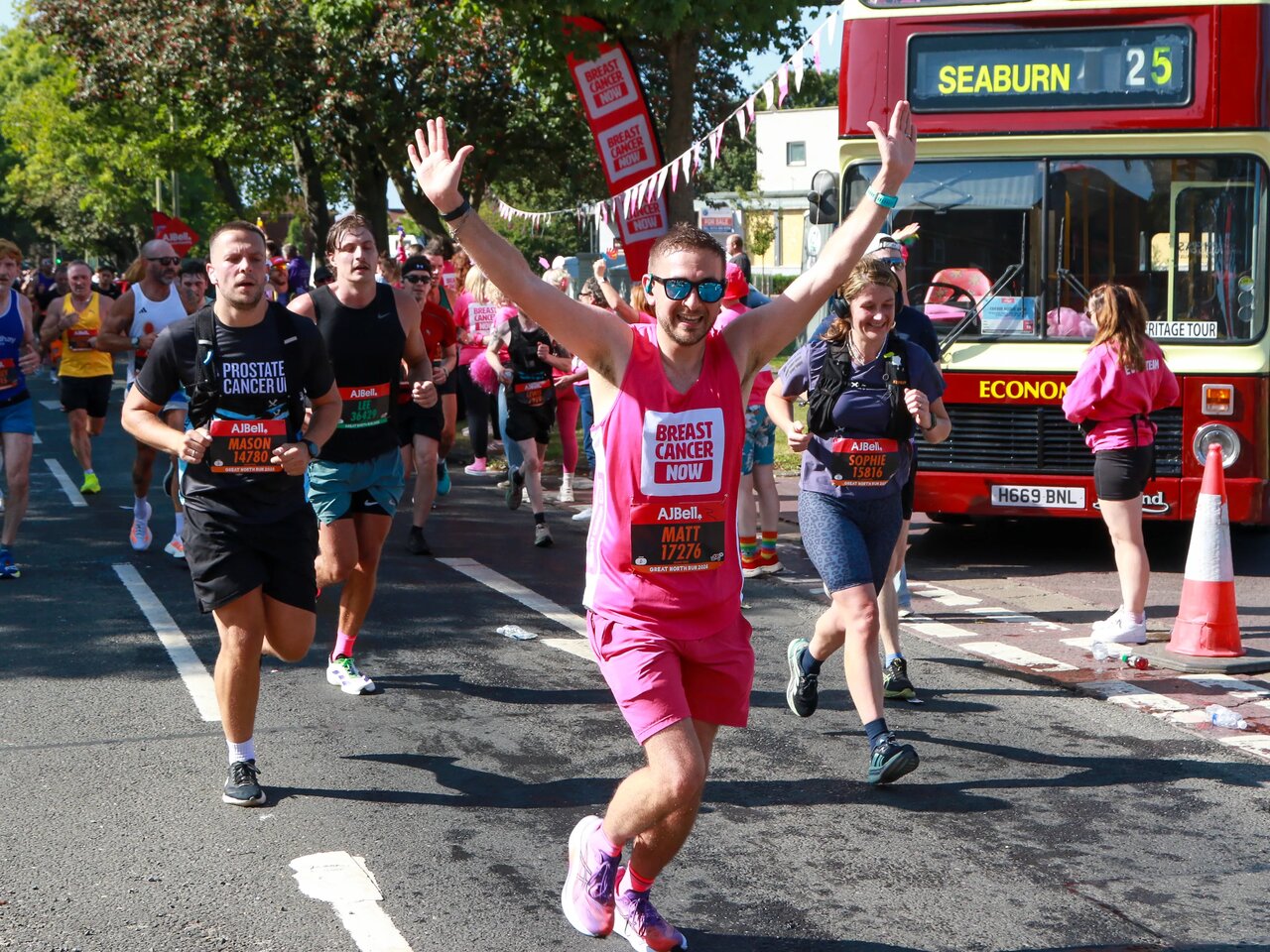 A man wearing a pink Breast Cancer Now running vest and pink shorts running in an event. There are other runners around him. He is holding is arms up and smiling at the camera.