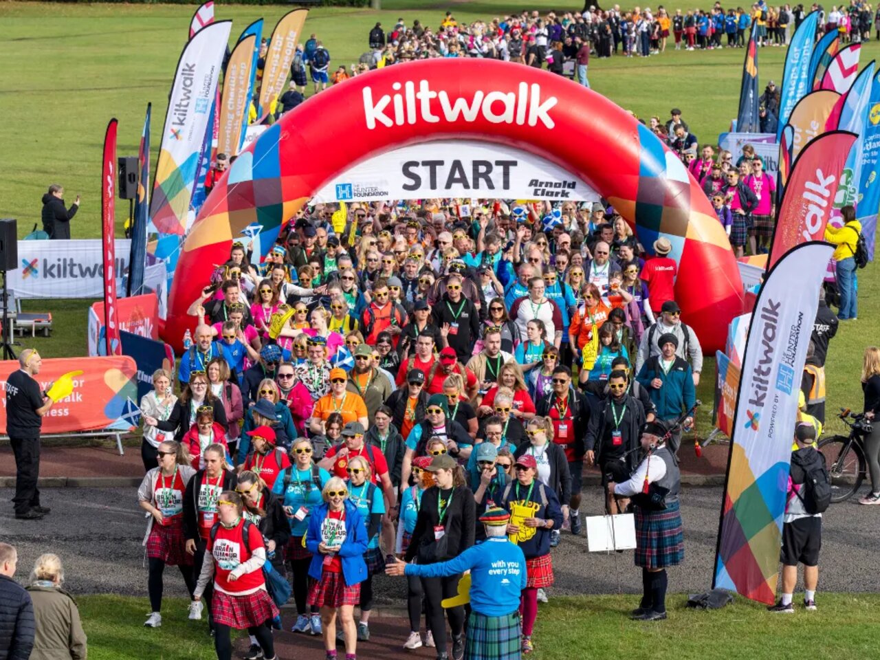 A crowd of participants passing under the inflatable arch at the start of Kiltwalk Edinburgh. They are wearing kilts and a man is playing the bagpipes.